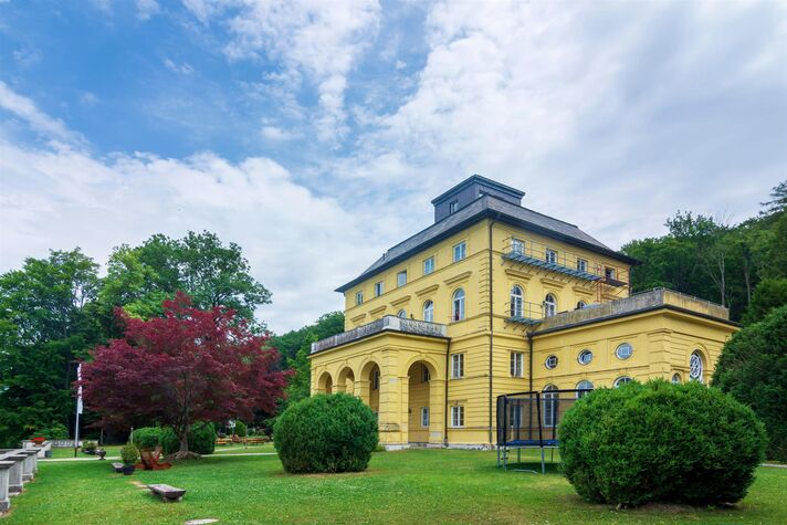 Gelb verputztes Schloss mit Säulenportal steht auf einer gepflegten Rasenfläche zwischen Buchsformschnitt und rot belaubtem Ahorn.