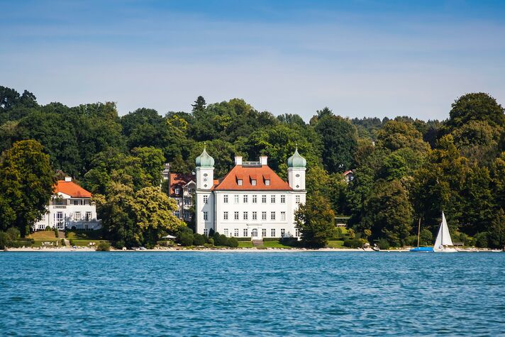 Weißes Schloss mit zwei grünen Zwiebeltürmen liegt dicht am Ufer, davor kreuzt ein Segelboot auf dem blauen See.