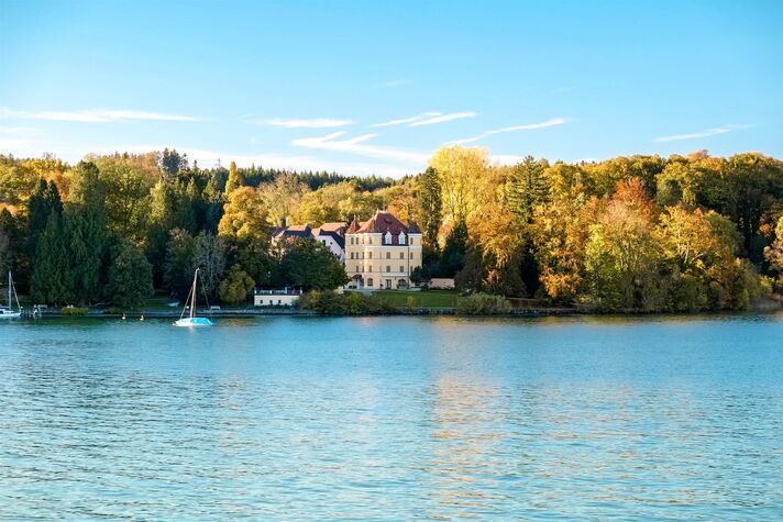 Vom Wasser aus gesehen steht ein helles Schloss auf einer bewaldeten Landzunge, davor ankert ein Segelboot.
