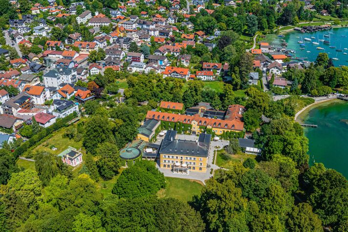 Luftaufnahme eines gelben Schlosses mit weitem Garten, dahinter die Dächer des Orts und der See.
