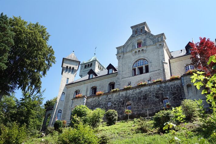 Massiver Bau im neuromanischen Stil mit hellem Rundturm und Zinnenkranz, umgeben von dichtem Grün.