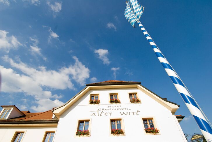 Hell verputzte Fassade des Hotels Alter Wirt mit geschwungenem Schriftzug, roten Blumenkästen an den Fenstern und einem blau-weißen Maibaum davor.