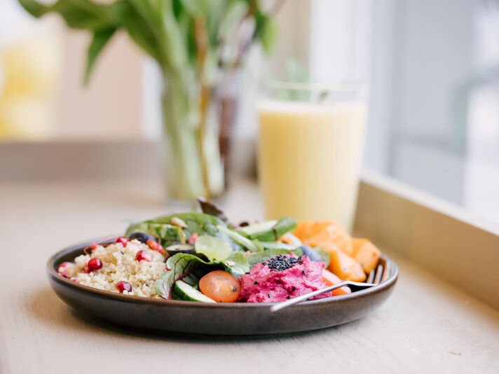 Dunkle Schale mit Couscous, Blattsalat, Roter Bete, Melone und Tomaten, dahinter ein hoher Becher mit hellgelbem Smoothie.
