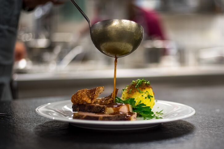 Aus einer Kelle fließt dunkle Sauce über ein Fleischgericht mit Kartoffelknödel, Kruste und Petersilie auf weißem Teller.