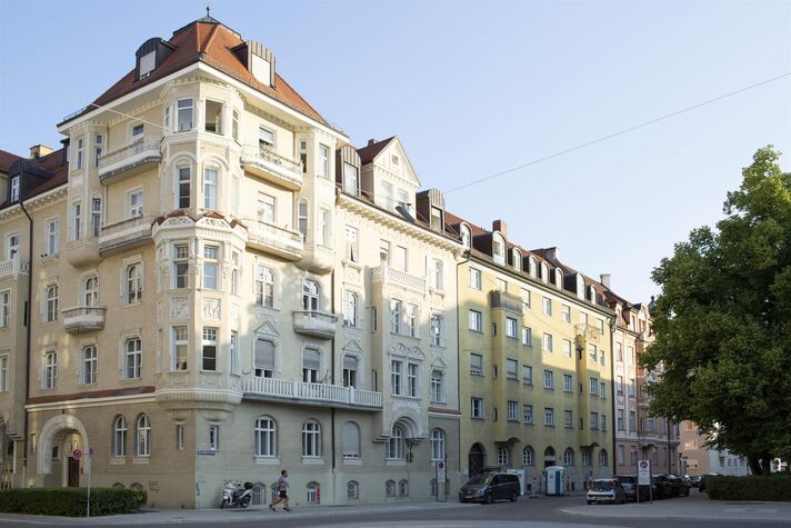 Straßenzug mit hellen Altbaufassaden in Ocker und Creme, Erkern, Stuckgesimsen und Balkonen, davor parkende Autos und Bäume.