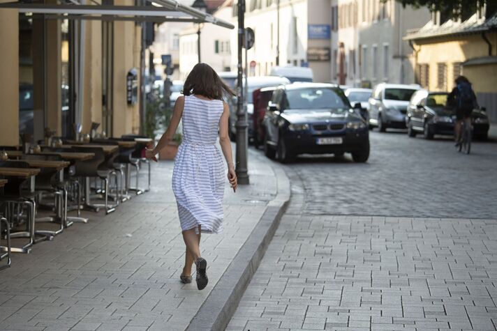 Eine Frau im gestreiften Sommerkleid geht über einen breiten Gehweg, links die bestuhlte Terrasse eines Cafés, rechts die Fahrbahn.
