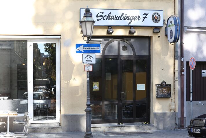 Eingang der Kneipe Schwabinger 7 mit geschwungenem Schriftzug über der Tür, Laterne und Straßenschild an der Hausecke.