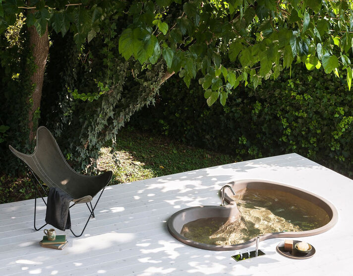 Ovale, in den Boden eingelassene Wanne mit Wasser und Armatur auf einer hellen Steinfläche, darüber überhängende Zweige mit Blättern.