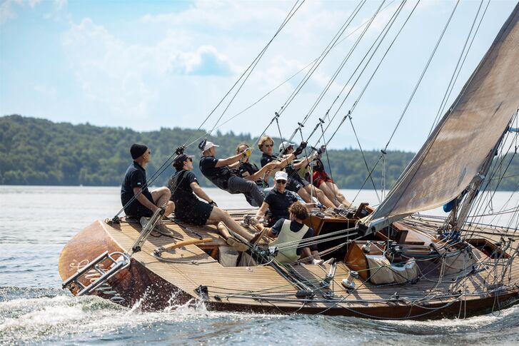 Eine hölzerne Yacht mit hohem Mast und heller Besegelung fährt mit mehreren Crewmitgliedern an Bord über den See, im Hintergrund bewaldete Hügel.