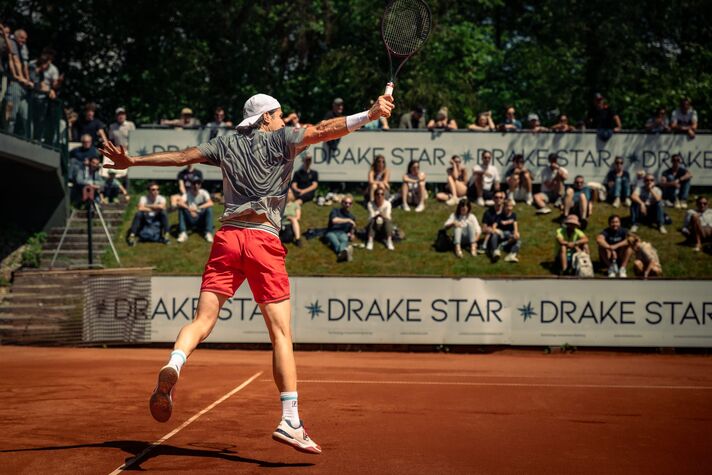 Ein Spieler schlägt auf dem Sandplatz eine gestreckte Vorhand, hinter der Bande sitzen und stehen Zuschauer im Sonnenlicht.