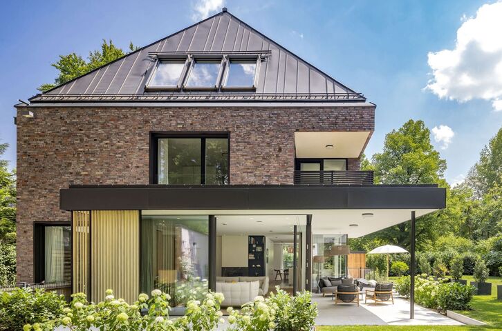 Neu gebaute Villa mit Ziegelfassade und Metalldach, im Erdgeschoss eine raumhohe Glasfront und eine überdachte Terrasse zum Garten.