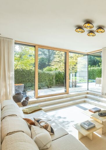 Heller Wohnraum mit tiefer Sitzlandschaft und Deckenleuchten, dahinter eine raumhohe Fensterfront mit Blick auf Terrasse und Hecke.