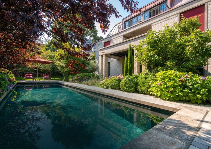 Langer Pool in einem eingewachsenen Garten, links Sonnenschirm und Liegestühle, rechts die Ziegelfassade einer Villa hinter blühenden Hortensien.