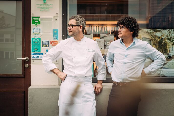 Zwei Männer in weißen Kochjacken stehen vor der grün gestrichenen Eingangstür eines Restaurants.