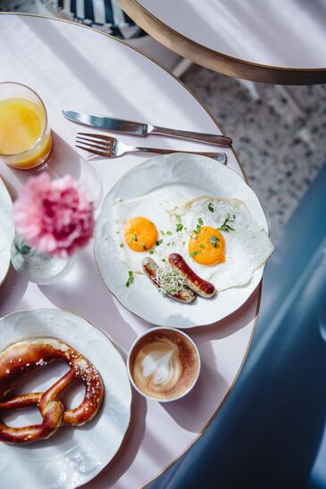 Von oben fotografierter Frühstückstisch mit Spiegeleiern, Brezn, Aufschnitt und Blumen zwischen den Tellern.