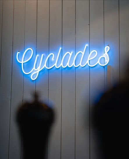 Blau leuchtender Schriftzug „Cyclades“ auf einer dunklen Holzwand neben einer schmalen Tür.