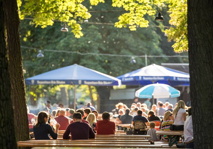 Dicht besetzte Biergartenbänke unter alten Kastanien, dazwischen aufgespannte blau-weiße Sonnenschirme.