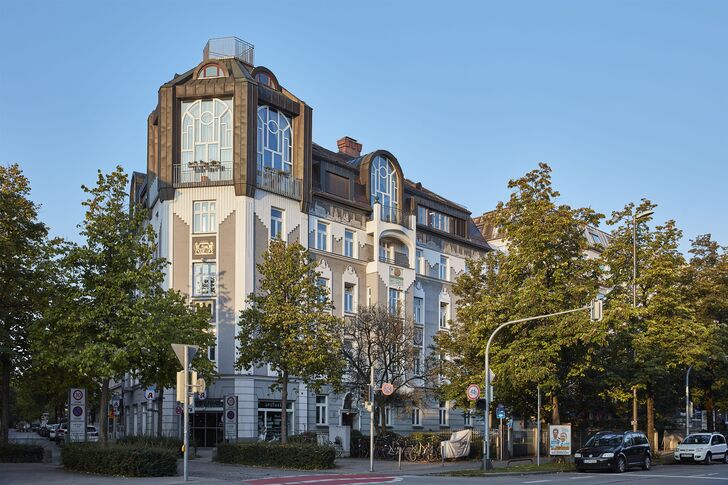 Mehrgeschossiges Eckhaus mit Erkerturm und Dachaufbau an einer von Bäumen gesäumten Straßenkreuzung.