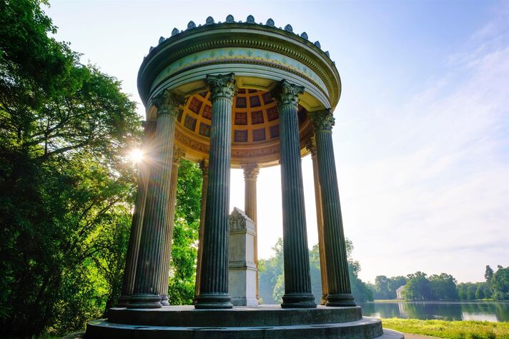 Runder Säulentempel mit grünem Kuppeldach steht im Gegenlicht zwischen hohen Bäumen am Seeufer.