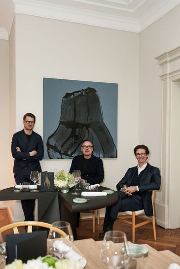 Three men at a laid table in front of a large painting at the launch of the event series in Munich.
