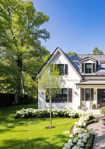 White country house with dormers and shutters behind a garden with flowering hydrangeas and mature trees.