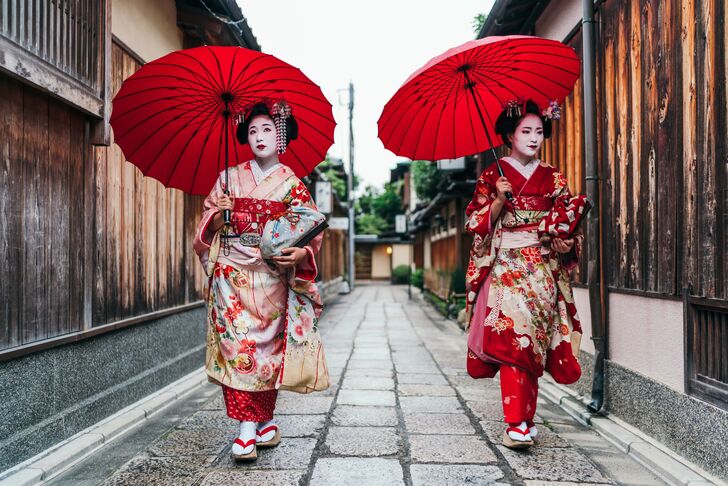 Zwei Frauen in Kimono mit roten Papierschirmen gehen eine schmale Gasse zwischen Holzhäusern entlang.