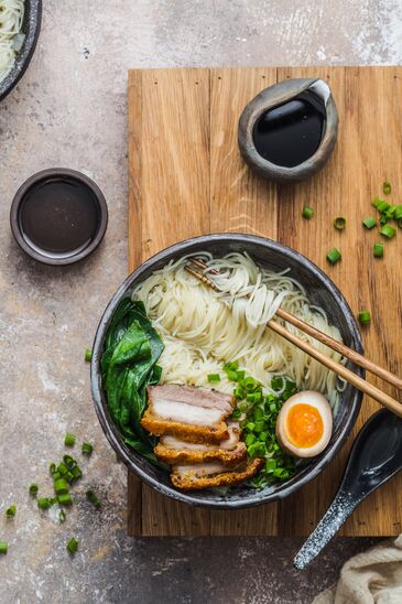 Von oben fotografierte Schale mit Nudelsuppe, Ei, Fleischscheiben und Frühlingszwiebeln auf hellem Holz.
