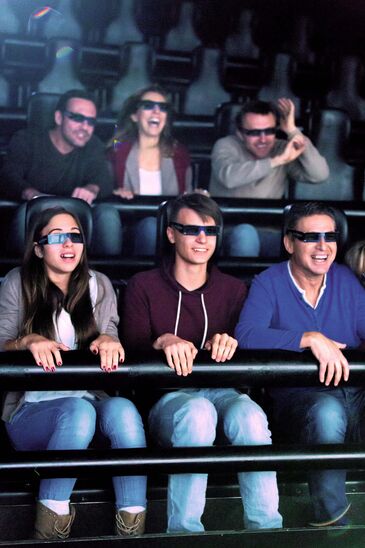 Kinobesucher in zwei Sitzreihen tragen 3D-Brillen und reagieren lachend und mit erhobenen Händen auf den Film.