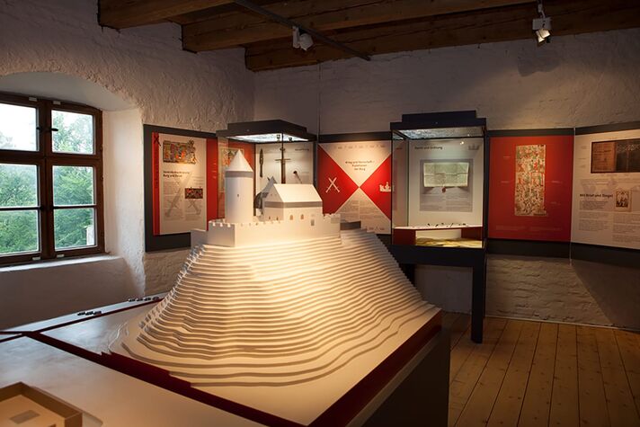 Ausstellungsraum mit weiß getünchten Wänden, in der Mitte ein großes Holzmodell einer Burganlage, dahinter Vitrinen und Schautafeln.