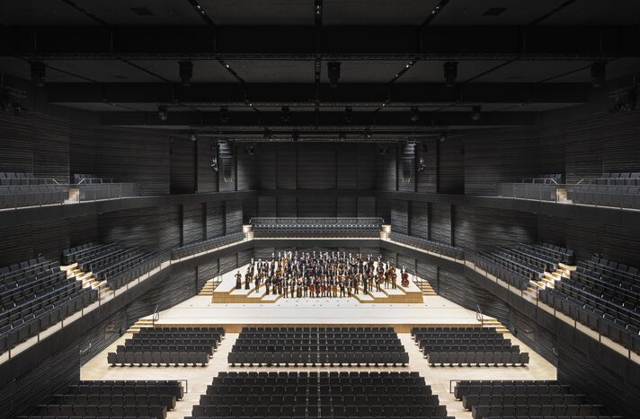Weiter Konzertsaal in dunklen Tönen mit heller Bühneninsel, auf der ein Orchester spielt, ringsum umlaufende Ränge.