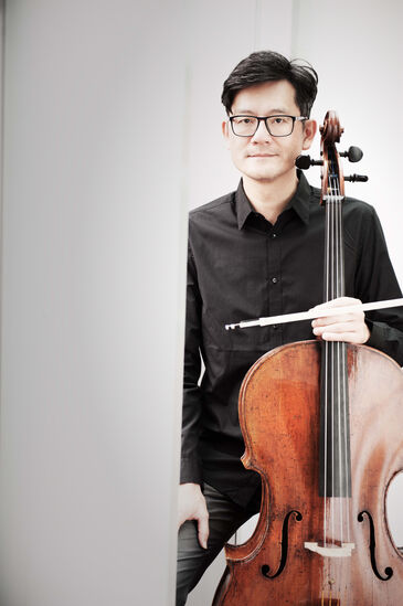 Der Cellist Wen-Sinn Yang steht im schwarzen Hemd vor hellgrauem Hintergrund, den Bogen in der Hand, das Cello an der Seite.
