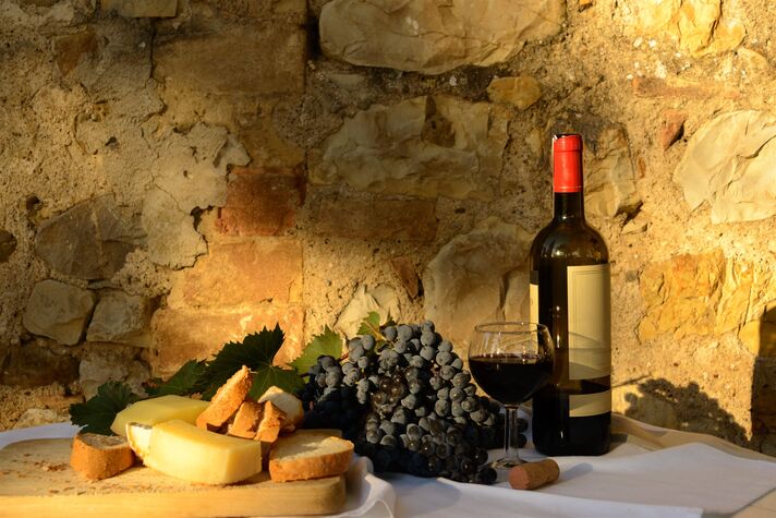 Rotweinflasche und Glas neben Pecorino-Stücken, Trauben und Brot auf weißem Tuch vor einer Tuffsteinwand.