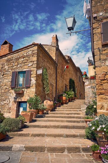 Schmale Gasse mit Natursteinhäusern, Blumentöpfen an den Wänden und einer Treppe, die zwischen den Fassaden aufsteigt.
