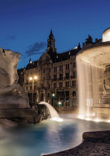 Beleuchteter Brunnen mit Steinfiguren und Wasserbecken in der Münchner Innenstadt vor einer Gründerzeitfassade zur blauen Stunde.
