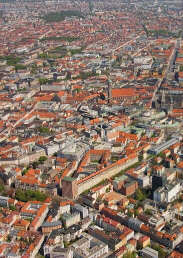Luftbild über das dicht bebaute Münchner Stadtgebiet mit den roten Ziegeldächern der Innenstadt.