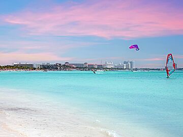 A tropical beach with a turquoise-blue sea, windsurfers, and a pink evening sky