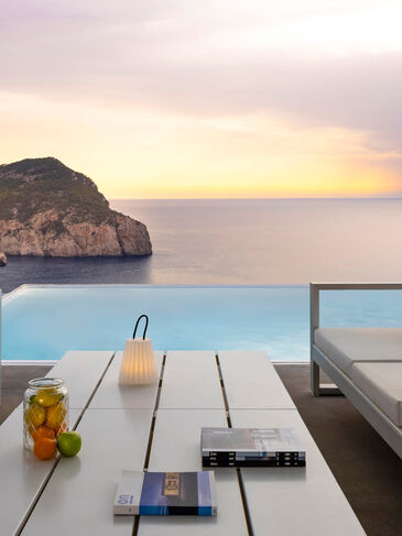 Überdachte Terrasse mit hellen Loungemöbeln vor einem Pool, dahinter Felsküste und Meer im Abendlicht
